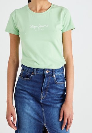 Kobieta w jasnozielonej koszulce Pepe Jeans London, wpuszczonej w niebieską dżinsową spódnicę z rozcięciem z przodu i zapięciem na guziki.