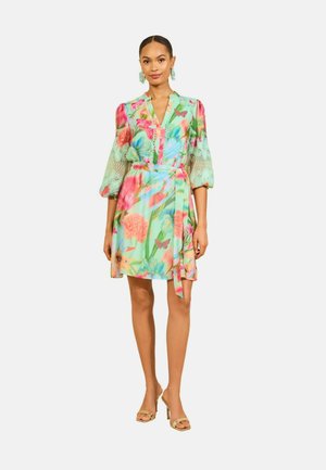 Femme portant une robe fleurie colorée avec des manches en dentelle bouffantes et une ceinture à la taille, associée à des sandales à talons beiges et des boucles d’oreilles assorties.