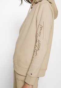 Beżowy hoodie wykonany z miękkiej tkaniny, z dużą kieszenią z przodu, kapturem ze sznurkiem oraz haftowanym logo na rękawie i z przodu.
