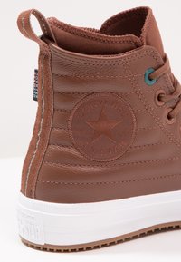 Hnědé kožené vysoké tenisky s texturovaným prošíváním, gumovou podrážkou a kulatou nášivkou s logem obsahující hvězdu a text "Converse All Star".