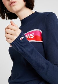 Mörkblå stickad tröja med en djärv färgblockgrafik i röd och rosa; ribbade ärmslut; logotypmärke på ärmen.