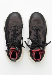 Mustat ja harmaat urheilukengät, joissa on teksturoitu päällinen, pyöreät nauhat, punaiset yksityiskohdat kauluksessa ja valkoinen kumipohja. Hintalaput kiinnitetty.