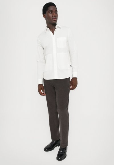 Uomo in piedi, che indossa una camicia bianca con bottoni e tre tasche frontali, pantaloni marroni scuri e scarpe in pelle nere, su uno sfondo bianco semplice.