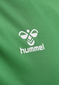 Wit gestileerd bijenlogo met chevronvormen en "hummel" tekst op een gladde groene stoffen achtergrond.