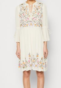 Vit klänning med blommig broderi i rosa, orange och grönt. Nyckelhålshalsringning med tofsar och trumpetärmar. Lätt, flytande tyg.