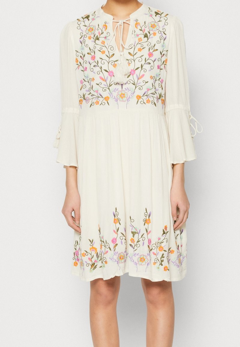Vit klänning med blommig broderi i rosa, orange och grönt. Nyckelhålshalsringning med tofsar och trumpetärmar. Lätt, flytande tyg.