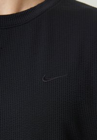 Svart strukturert langermet skjorte med rund hals og en subtil svart Nike-logo på brystet. Forsterket søm langs kantene.