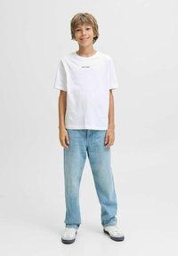 Chlapec stojící a usmívající se, oblečený v bílém tričku s malým textem, světle modrých džínách a bílých teniskách na čistém bílém pozadí.