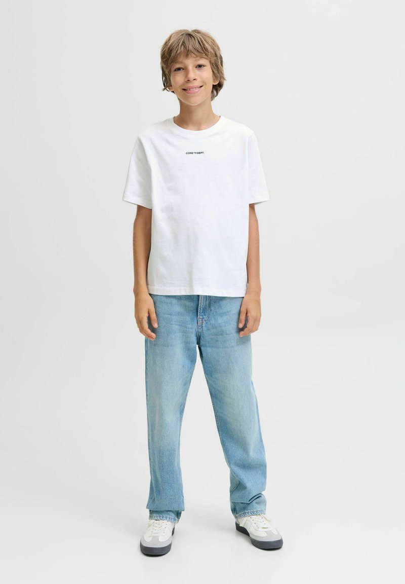 Chlapec stojící a usmívající se, oblečený v bílém tričku s malým textem, světle modrých džínách a bílých teniskách na čistém bílém pozadí.