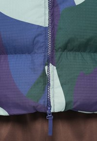 Close-up de uma jaqueta acolchoada multicolorida com um fecho azul sobre um fundo de tecido castanho.