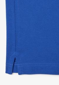 Tissu bleu texturé avec couture latérale et petite fente à l’ourlet, montrant un gros plan du bord du vêtement.