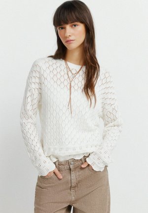 Młoda kobieta w białym swetrze z teksturowanego dziania i beżowych spodniach z wysokim stanem, z rękami w kieszeniach, stoi na gładkim tle.