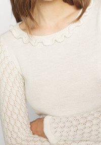 Sweter w kolorze kremowym o prążkowanej fakturze, z falbankowym dekoltem i koronkowymi, długimi rękawami. Subtelny i delikatny wzór.