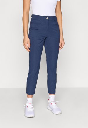 Frau trägt dunkelblaue, enganliegende, gekürzte Hose, weiße Knöchelsocken, weiße Sportschuhe und ein weißes kurzärmeliges Oberteil, steht auf einem hellen Boden.
