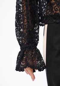 Ręka w czarnej koronkowej widoczkowej rękawie z marszczonym mankietem, obok czarnych i beżowych spodni w pionowe paski na białym tle.