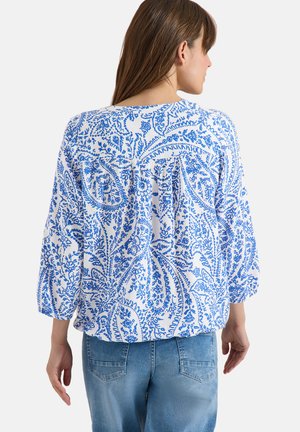 Femme portant un chemisier blanc avec un imprimé floral paisley bleu et un jean bleu, vue de dos sur un fond uni.