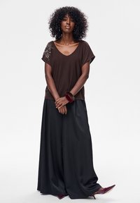 Brązowa bluzka z dekoltem w serek i krótkimi rękawami, ozdobiona srebrnymi zdobieniami na jednym ramieniu, w połączeniu z czarnymi spodniami o szerokich nogawkach i bordowymi szpilkami.