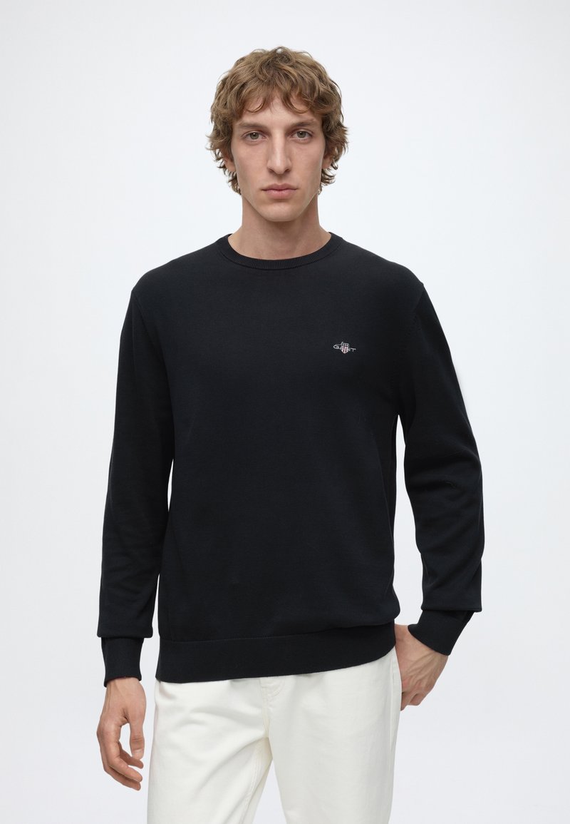 Young man with short curly hair wears black long-sleeve sweatshirt and white pants, standing against a plain white background.