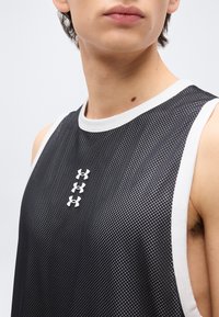 Κοντινό πλάνο ατόμου που φοράει μαύρο αμάνικο αθλητικό πουκάμισο από δίχτυ με τρία λευκά λογότυπα Under Armour στο κέντρο του στήθους.