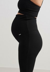 Svart ribbet graviditetsleggings med høy midje og en jevn tekstur. Liten metallisk detalj ved hoften. Fittet design.