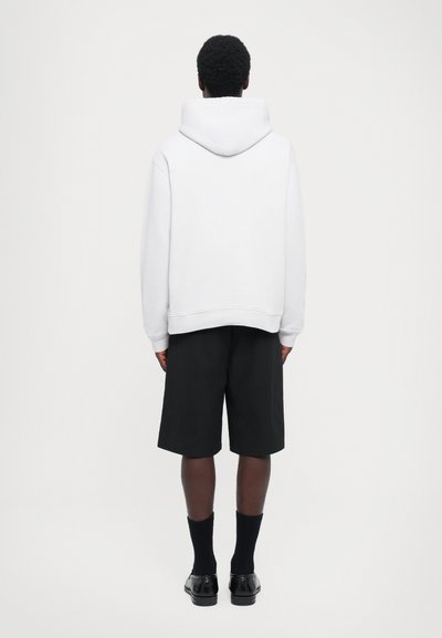 Person, der von hinten betrachtet wird, trägt einen weißen Kapuzenpullover, schwarze knielange Shorts, schwarze Socken und schwarze Schuhe vor einem weißen Hintergrund.