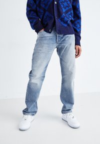 Hellblaue Jeans mit lockerer Passform, leicht verwaschen, kombiniert mit einem gemusterten dunkelblauen Cardigan und weißen Sneakers.