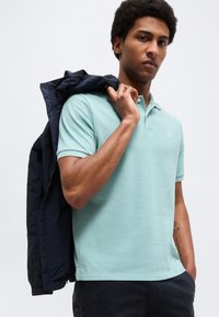 Muž v svetlomodrom tričku s golierom a tmavých nohaviciach, držiaci tmavú bundu prehodenú cez rameno na jednoduchom svetlom pozadí.