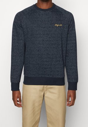 Mand iført navyblå mønstret langærmet sweater med gult "Originals"-tekst på brystet og beige bukser, stående mod en hvid baggrund.