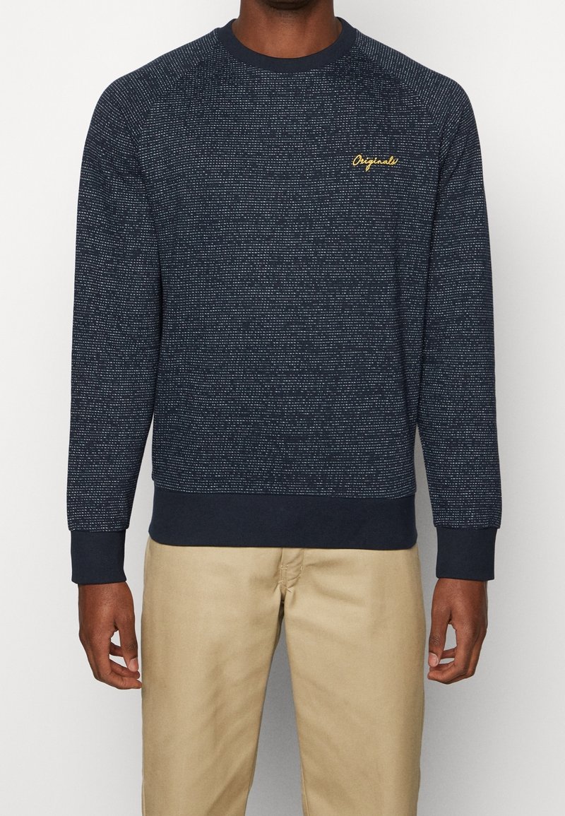 Homme portant un pull à manches longues bleu marine à motifs avec le texte jaune « Originals » sur la poitrine et un pantalon beige, debout devant un fond blanc.