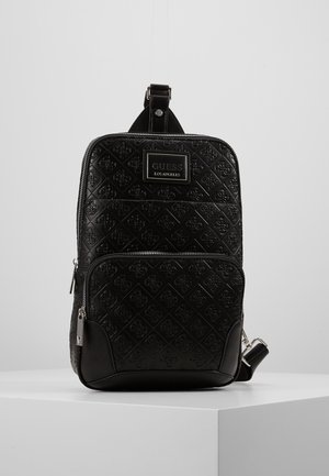 Sac à dos bandoulière noir GUESS avec motif de logo embossé, poche avant zippée et sangle d’épaule réglable, exposé sur un socle blanc.