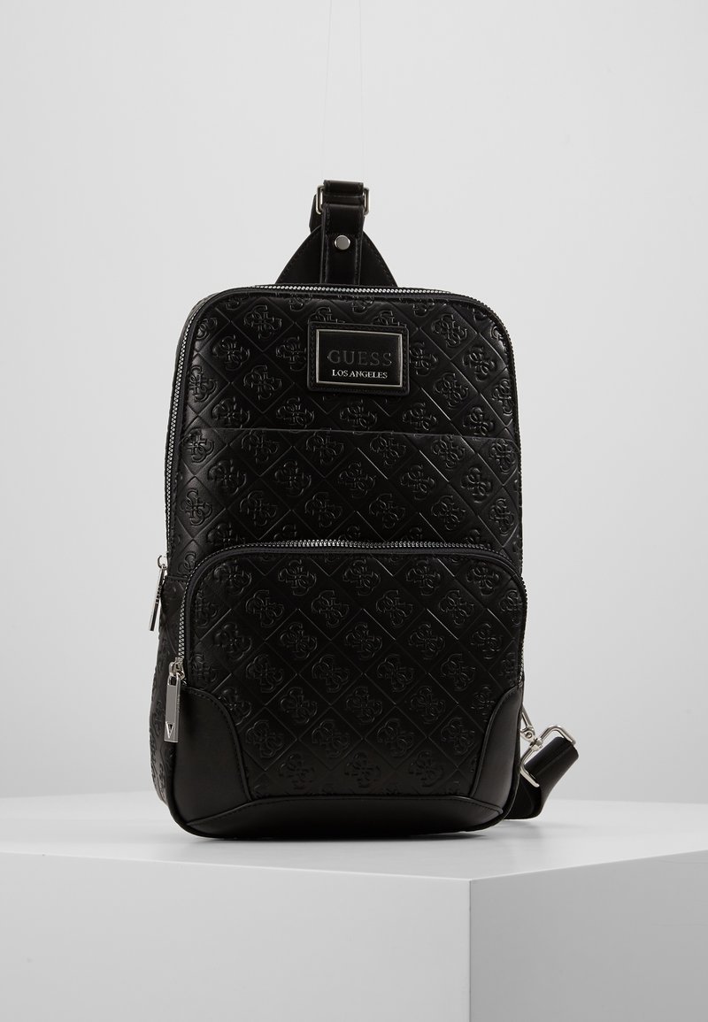 Sac à dos bandoulière noir GUESS avec motif de logo embossé, poche avant zippée et sangle d’épaule réglable, exposé sur un socle blanc.