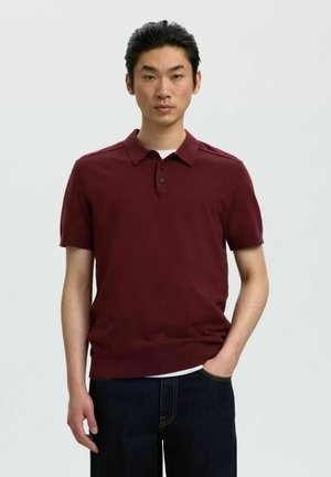 Hombre con una camiseta polo de manga corta color burdeos y vaqueros oscuros, de pie con una mano en el bolsillo frente a un fondo liso.
