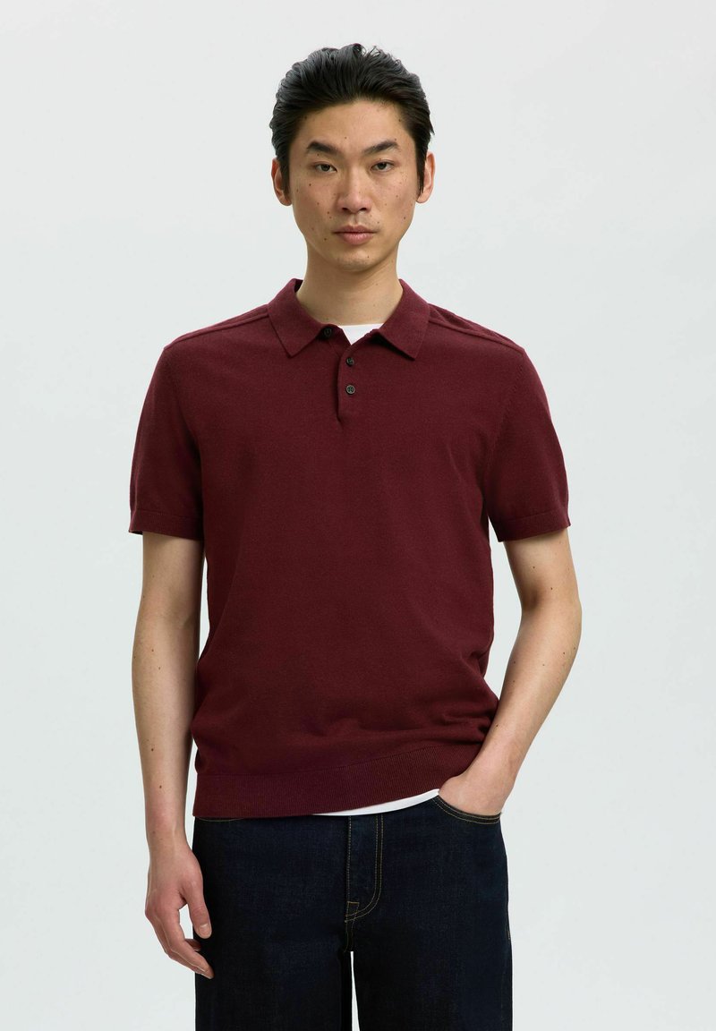 Homme portant un polo bordeaux à manches courtes et un jean foncé, debout avec une main dans la poche sur un fond uni.