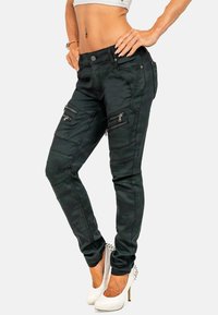 Pantaloni skinny camouflage verdi con molte tasche zip, caratterizzati da una texture liscia e un design aderente. Abbinati a scarpe con tacco alto bianche.