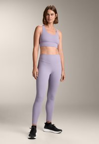 Lila športna modrček in hlače, izdelane iz elastičnega materiala, z gladko površino in prilegajočim dizajnom. Črne športne čevlje dopolnjujejo videz.