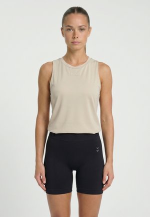 Vrouw met naar achteren gebonden haar, gekleed in een beige mouwloos sporttopje en zwarte aansluitende fietsshorts, staand tegen een effen witte achtergrond.