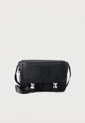 Sac messager noir Lacoste avec bandoulière ajustable, rabat avant, fermetures à boucle argentée et patch logo embossé.