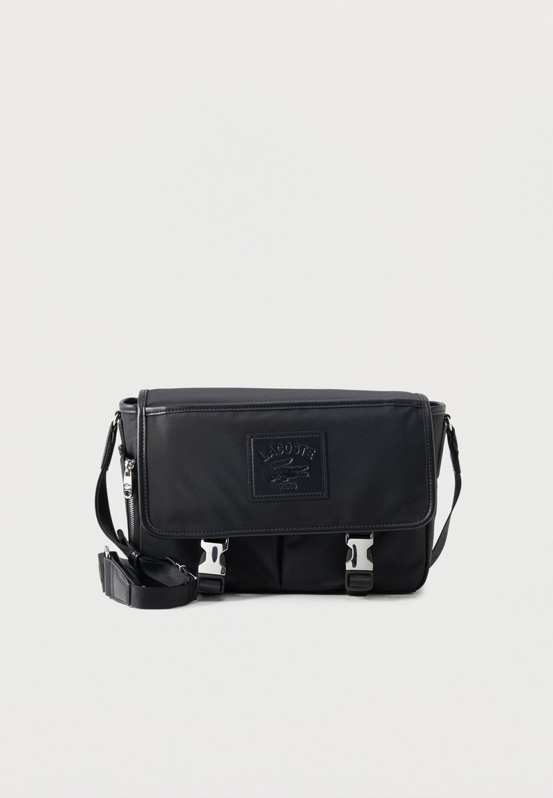 Μαύρη τσάντα ταχυδρόμου Lacoste με ρυθμιζόμενο ιμάντα ώμου, μπροστινό καπάκι, ασημί πόρπες και ανάγλυφο λογότυπο.