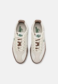 Sneakers Lacoste in tela color crema con dettagli marroni, presentano punta arrotondata, design con lacci e logo verde sul lato.
