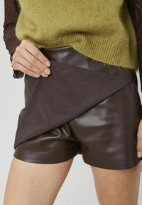 Shorts en similicuir marron avec un voile de tissu foncé drapé, associés à un pull en maille duveteuse vert clair. Texture lisse, coupe ajustée.