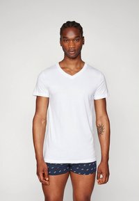 White cotton V-neck t-shirt on a male model. Features short sleeves, a relaxed fit, and a smooth texture. Model wears patterned navy shorts.