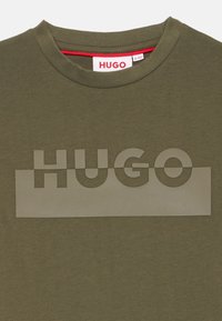 Alyvinės spalvos marškinėliai su apvalia apykakle, ant priekio puikuojasi didelis iškilus "HUGO" logotipas šviesesnės alyvinės spalvos.