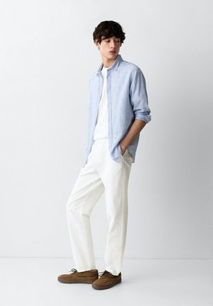 Jeune homme portant une chemise bleue claire boutonnée sur un t-shirt blanc, un pantalon blanc et des chaussures marron, debout les mains dans les poches devant un fond uni.