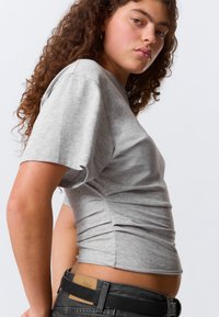 Cămașă gri cu mâneci scurte din material moale; are un design mulat cu detaliu adunat pe lateral. Asortată cu blugi închiși la culoare și o curea neagră.