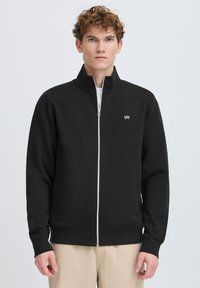Sweat-shirt zippé noir avec un col montant, des poignets et une ceinture côtelés. Présente un petit logo blanc sur le côté gauche de la poitrine. Texture de tissu lisse.