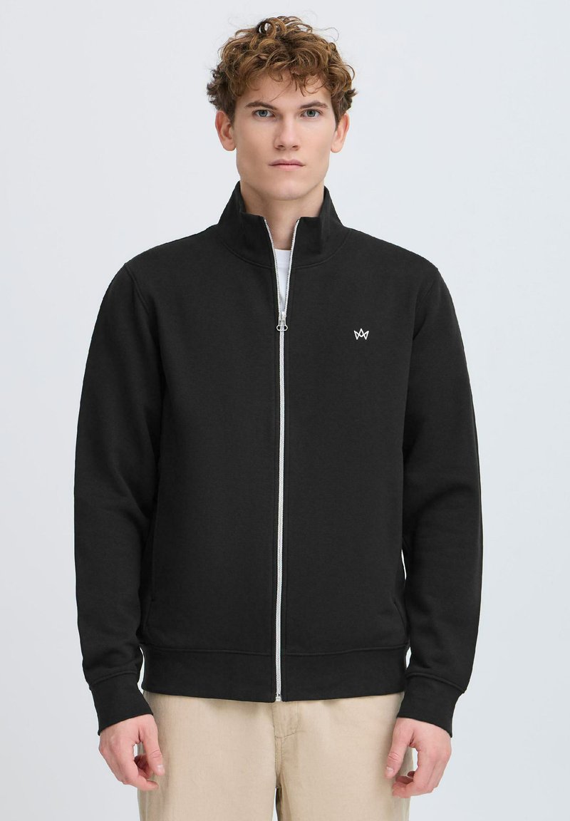 Sweat-shirt zippé noir avec un col montant, des poignets et une ceinture côtelés. Présente un petit logo blanc sur le côté gauche de la poitrine. Texture de tissu lisse.