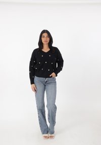 Schwarzer V-Ausschnitt-Pullover mit goldenen herzförmigen Verzierungen, kombiniert mit hellblauen Schlagjeans und offenen weißen Schuhen.