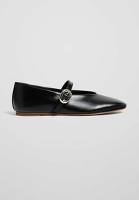 Ballerine plate en cuir noir avec une bride sur le cou-de-pied, une boucle dorée et une semelle fine et plate, présentée de côté sur fond uni.