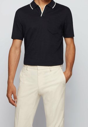 Mann in schwarzem Poloshirt mit kurzen Ärmeln und weißem Saum, mit cremefarbener Hose, eine Hand in der Tasche, vor einem einfarbigen Hintergrund.