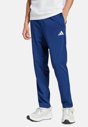 Man draagt blauwe Adidas trainingsbroek, witte sneakers en een wit shirt, staat met één hand in de zak tegen een effen achtergrond.